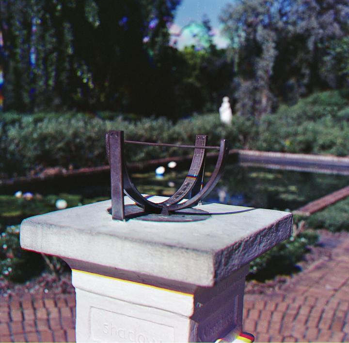 A sundial sits atop a stone pedestal in a garden with a pond and lush greenery in the background.