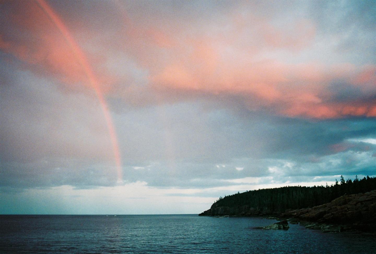 A rainbow stretches across a cloudy sky with pink and blue hues over a body of water and a tree-lined coast.
