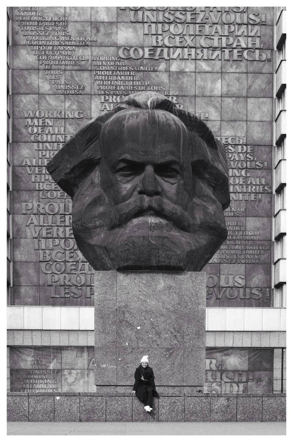 A woman sits on a ledge in front of a large statue of Karl Marx, with text carved into the wall behind it.