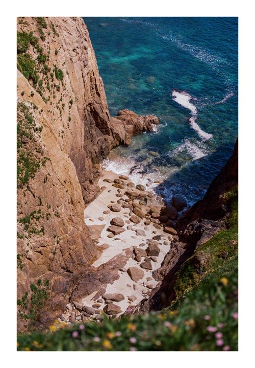 A hidden cove with a sandy beach and clear blue water is nestled between steep cliffs.