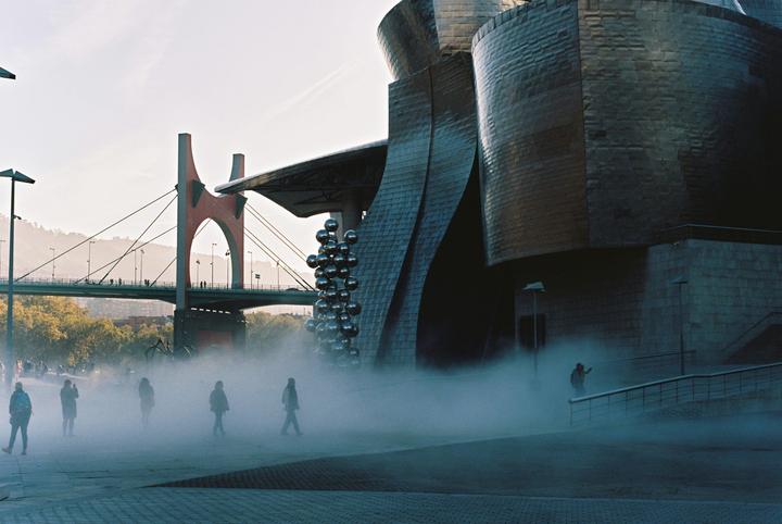 People walk through a misty plaza in front of a modern building and a bridge.
