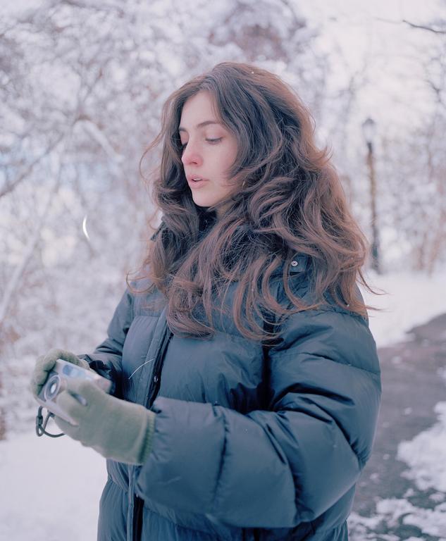 A woman wearing a puffer jacket and gloves holds a camera in a snowy landscape.