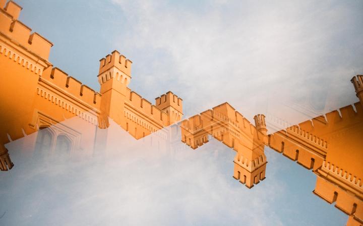 A double exposure shows the orange facade of a building with battlements against a blue sky.