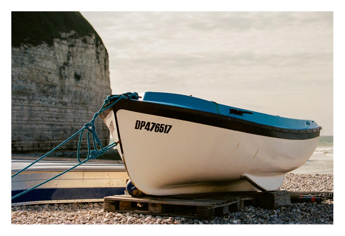 A white and blue boat sits on a rocky beach with a large cliff in the background.