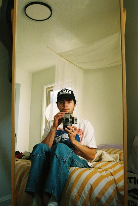 A man wearing a baseball cap and jeans sits on a bed and takes a mirror self-portrait with a camera.