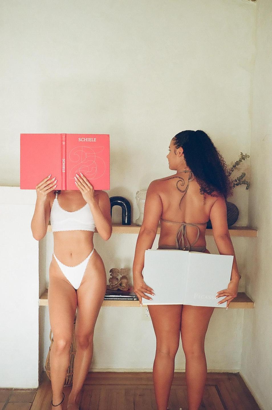 Two women stand in a room, one holding a red book to her face and the other holding a white book behind her.