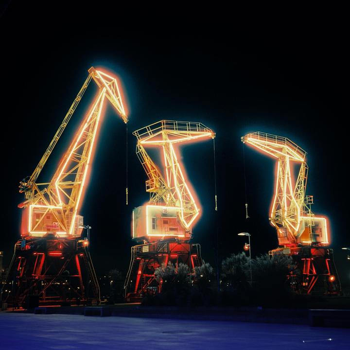 Three large cranes are illuminated with neon lights against a dark night sky.