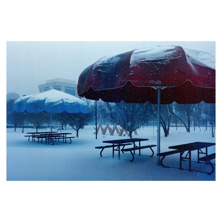 A snow-covered park with picnic tables and large umbrellas is seen under a hazy sky.