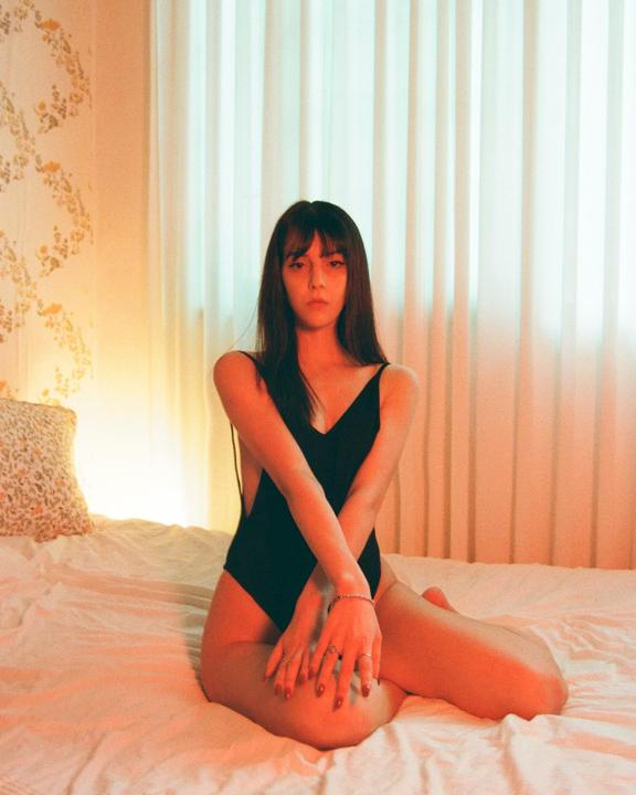 A woman in a black swimsuit sits on a bed with her arms crossed, looking directly at the viewer.