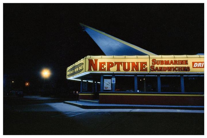 A brightly lit Neptune Submarine Sandwiches restaurant stands out against a dark night sky.
