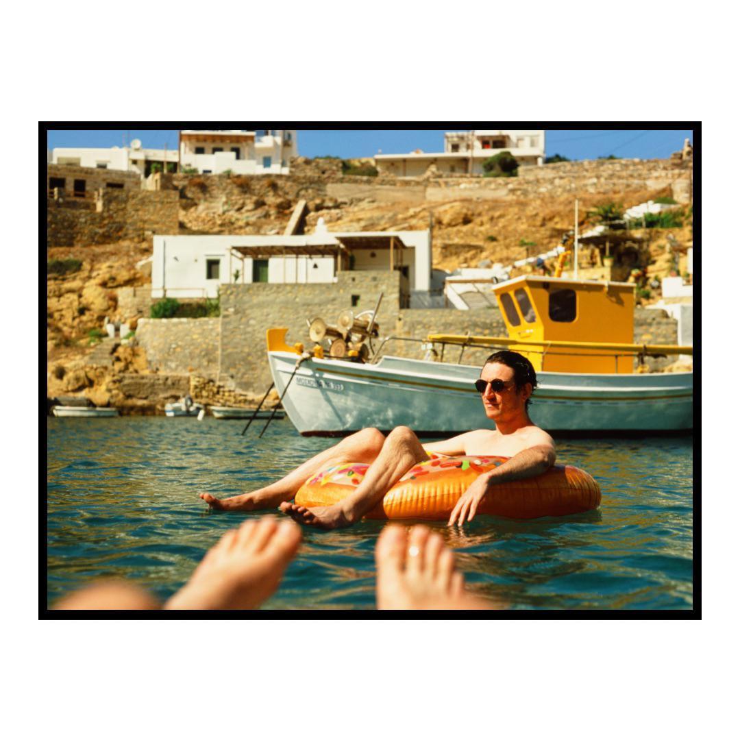 A man floats on an inflatable ring in the sea, with a village and boats on the shore in the background.