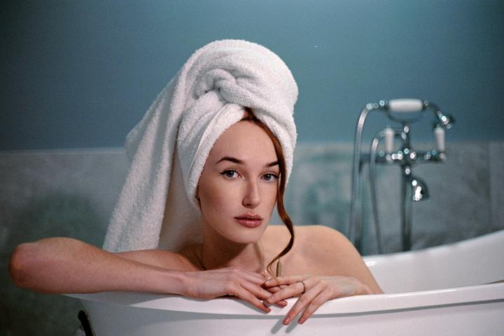 A woman with a towel wrapped around her head is in a bathtub, resting her arms on the edge.