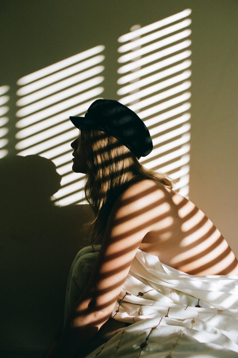 A woman wearing a hat is illuminated by striped shadows from a window blind.