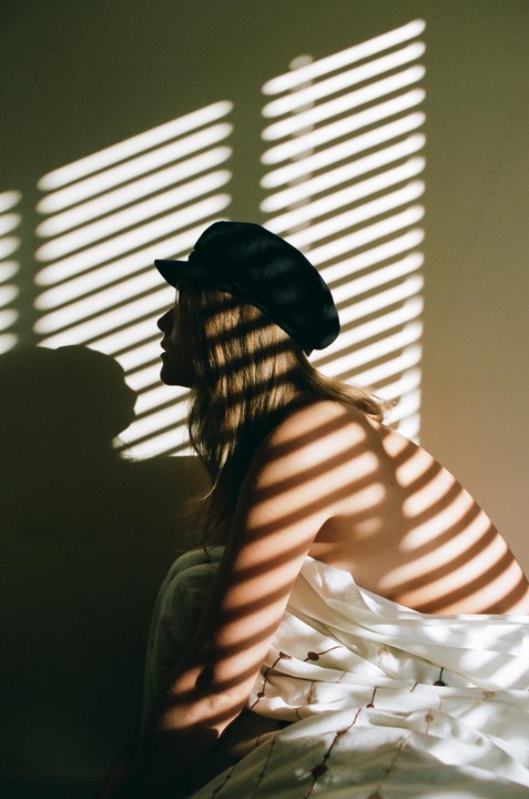 A woman wearing a hat is illuminated by striped shadows from a window blind.