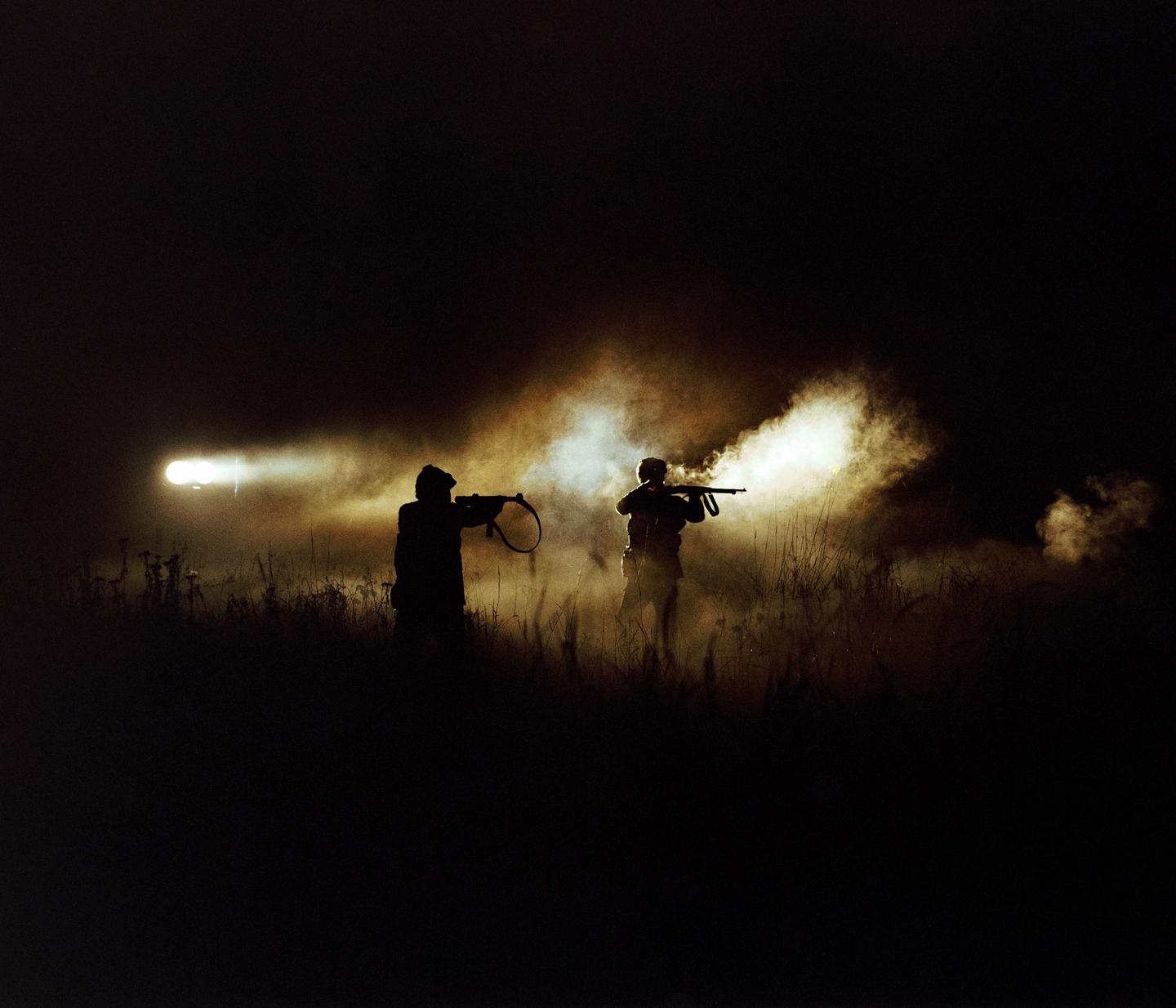 Two men are silhouetted against a bright light and smoke in a field at night.