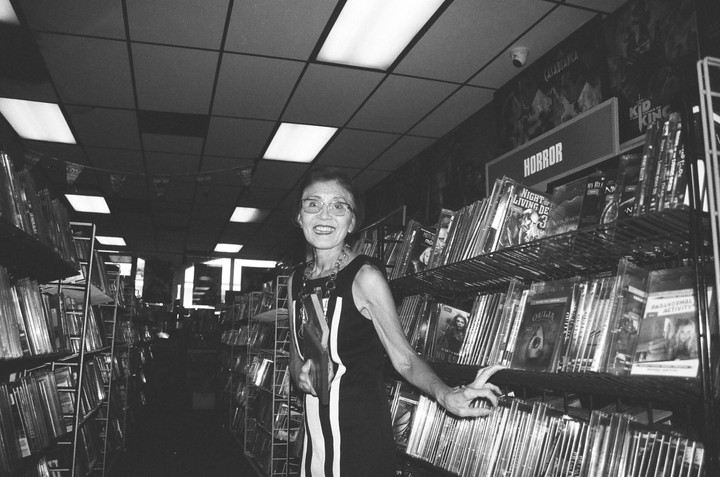 A smiling woman stands in a video store aisle next to shelves of horror movies.