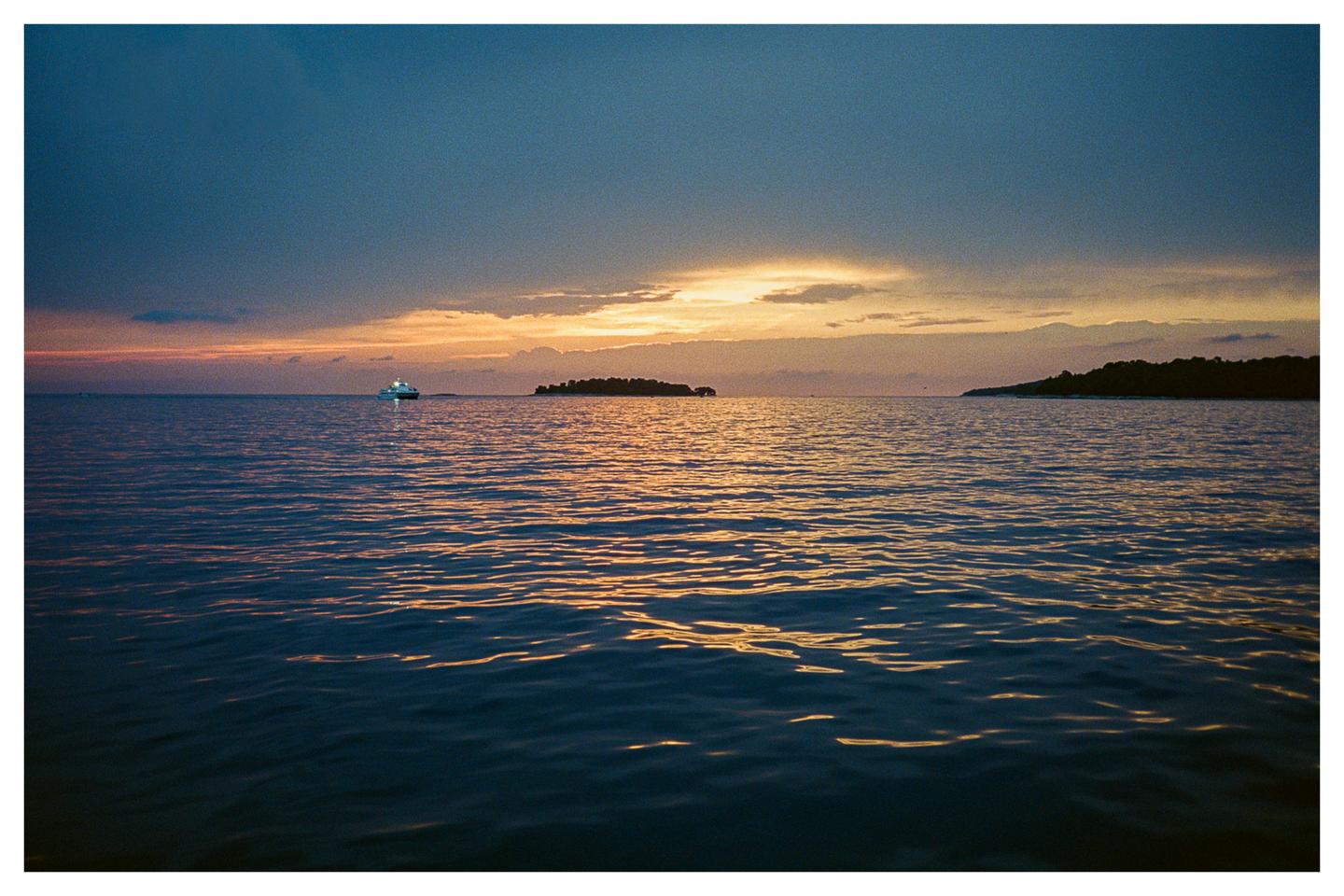 A boat with lights on is visible on the water with islands in the distance under a dramatic sunset sky.