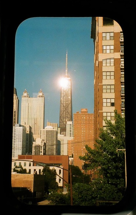 A cityscape with a prominent skyscraper reflecting sunlight is viewed through a train window.