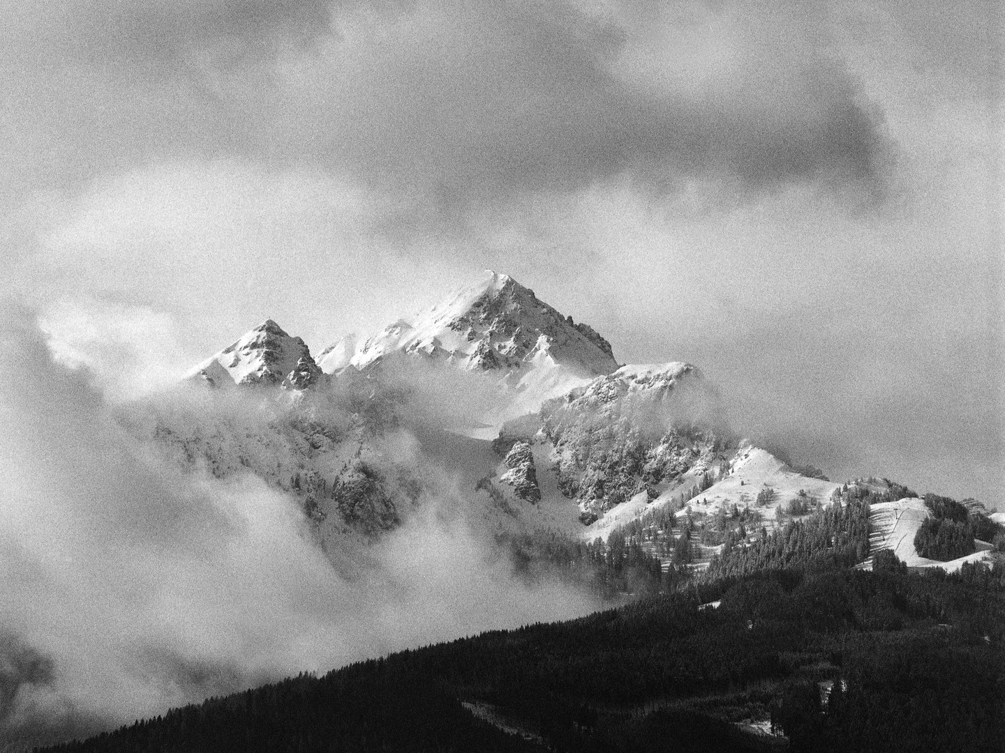 Snow-covered mountains are partially obscured by clouds, with a dark forest in the foreground.