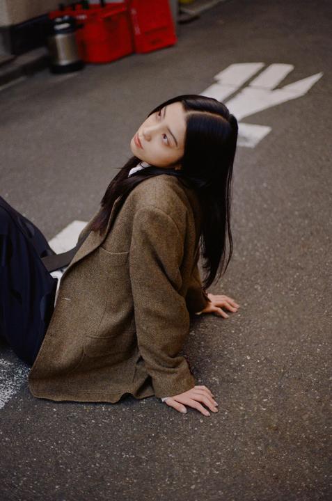 A woman in a brown jacket sits on an asphalt street with white markings.
