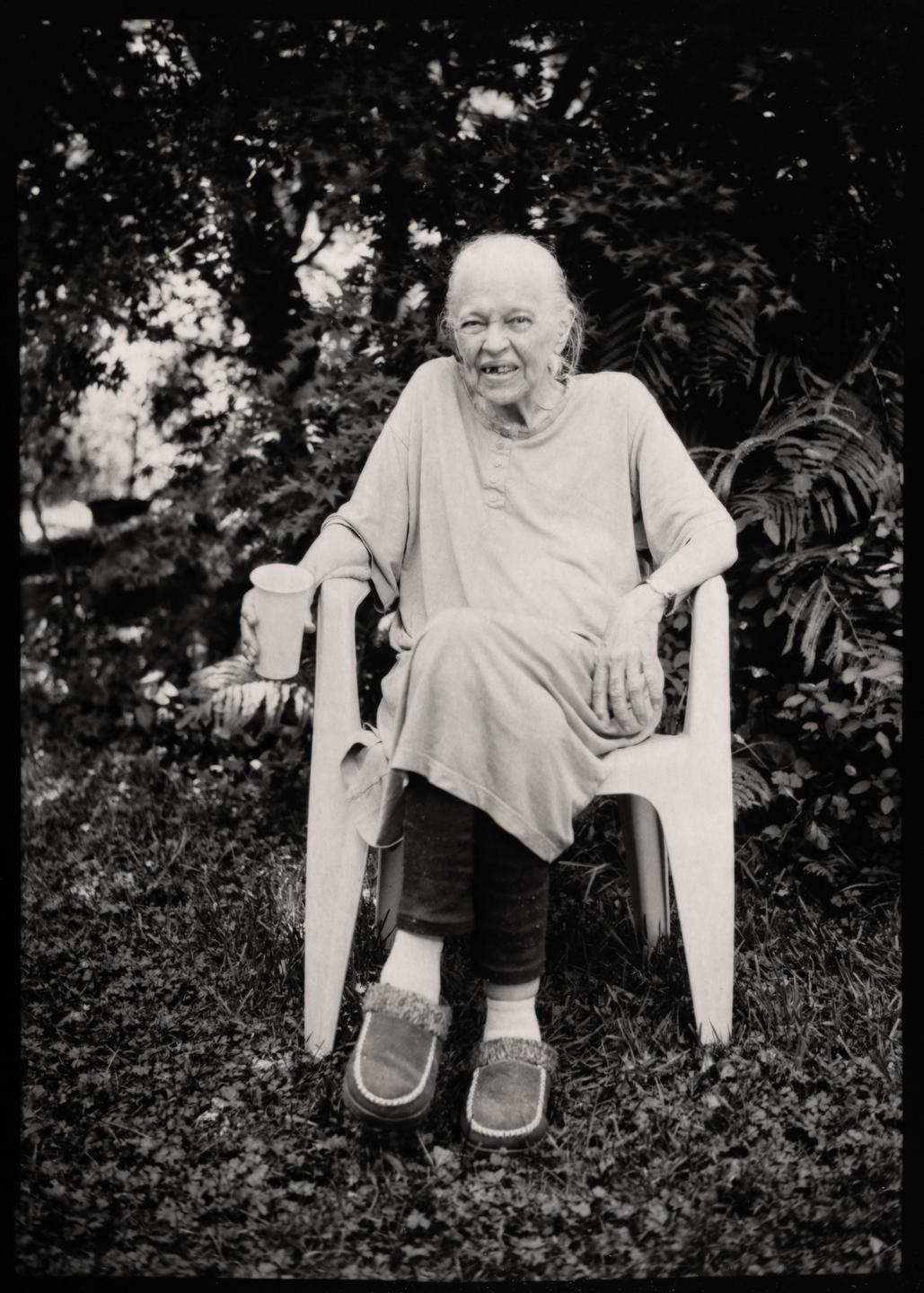 An elderly woman with white hair and a missing front tooth sits in a chair outdoors, holding a cup.