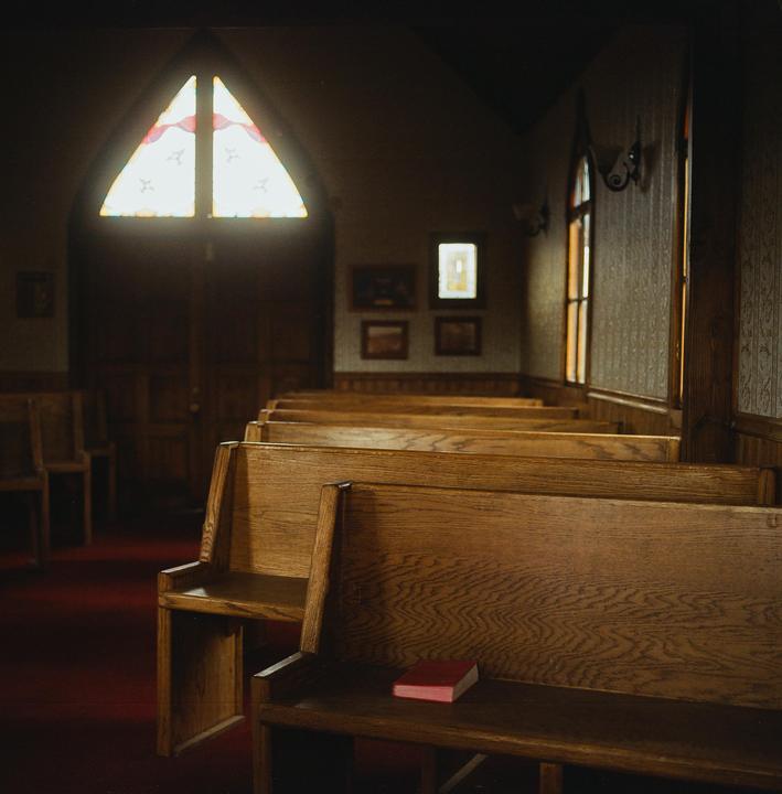 A red book rests on a wooden pew inside a church with stained glass windows.