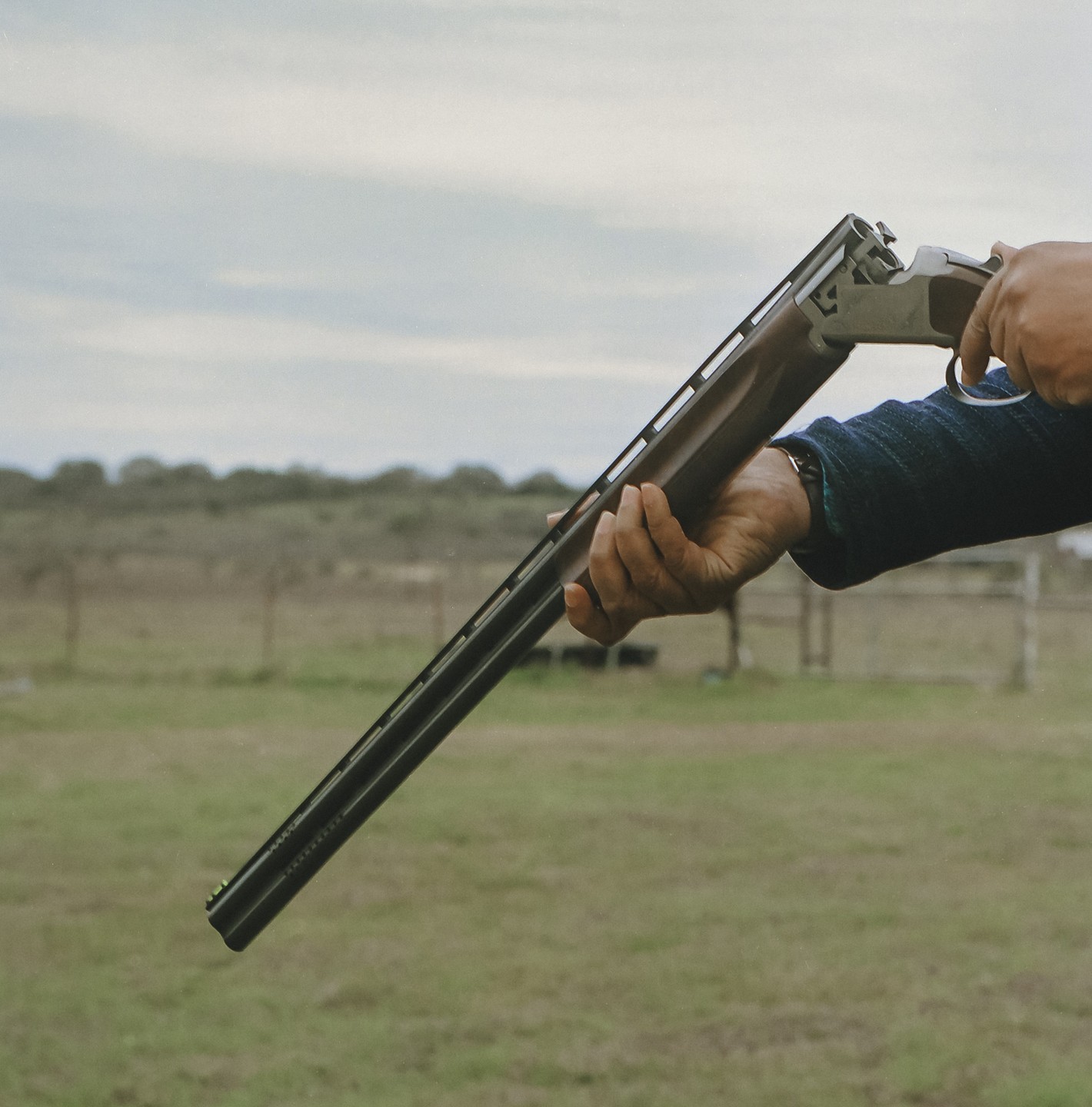 A person's hands are holding a shotgun in an open field under a cloudy sky.