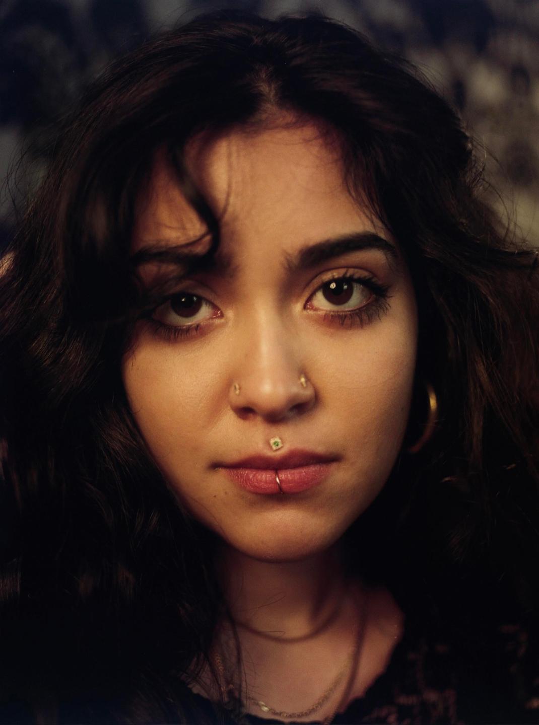 A woman with dark, curly hair and multiple facial piercings looks directly at the viewer.