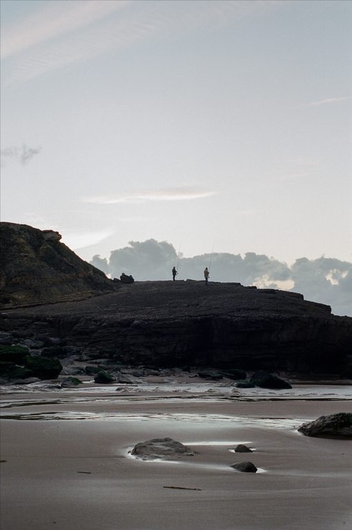 Two men fish from a rocky outcrop overlooking a sandy beach with tidal pools.
