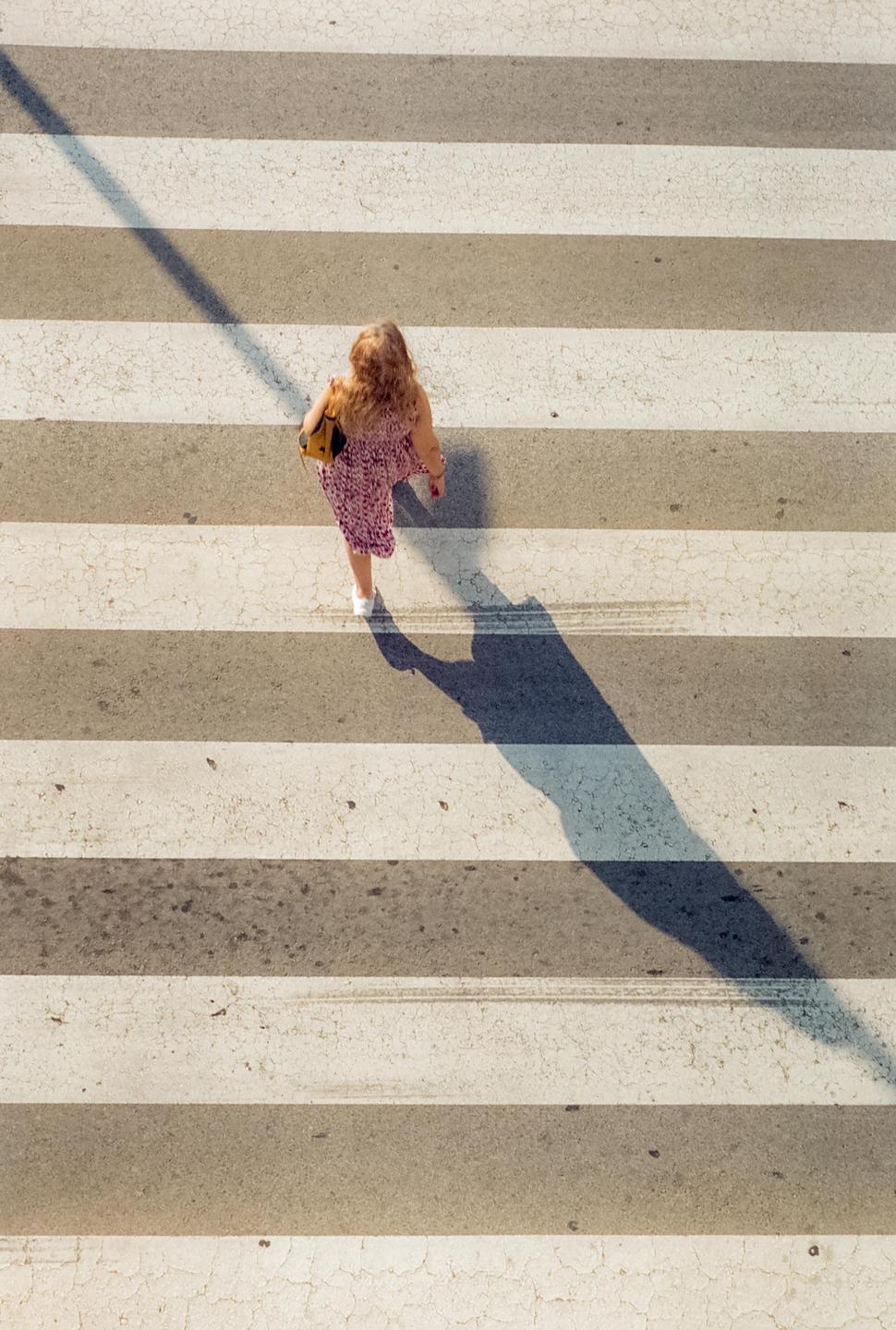 A woman in a patterned dress walks across a zebra crossing, casting a long shadow.
