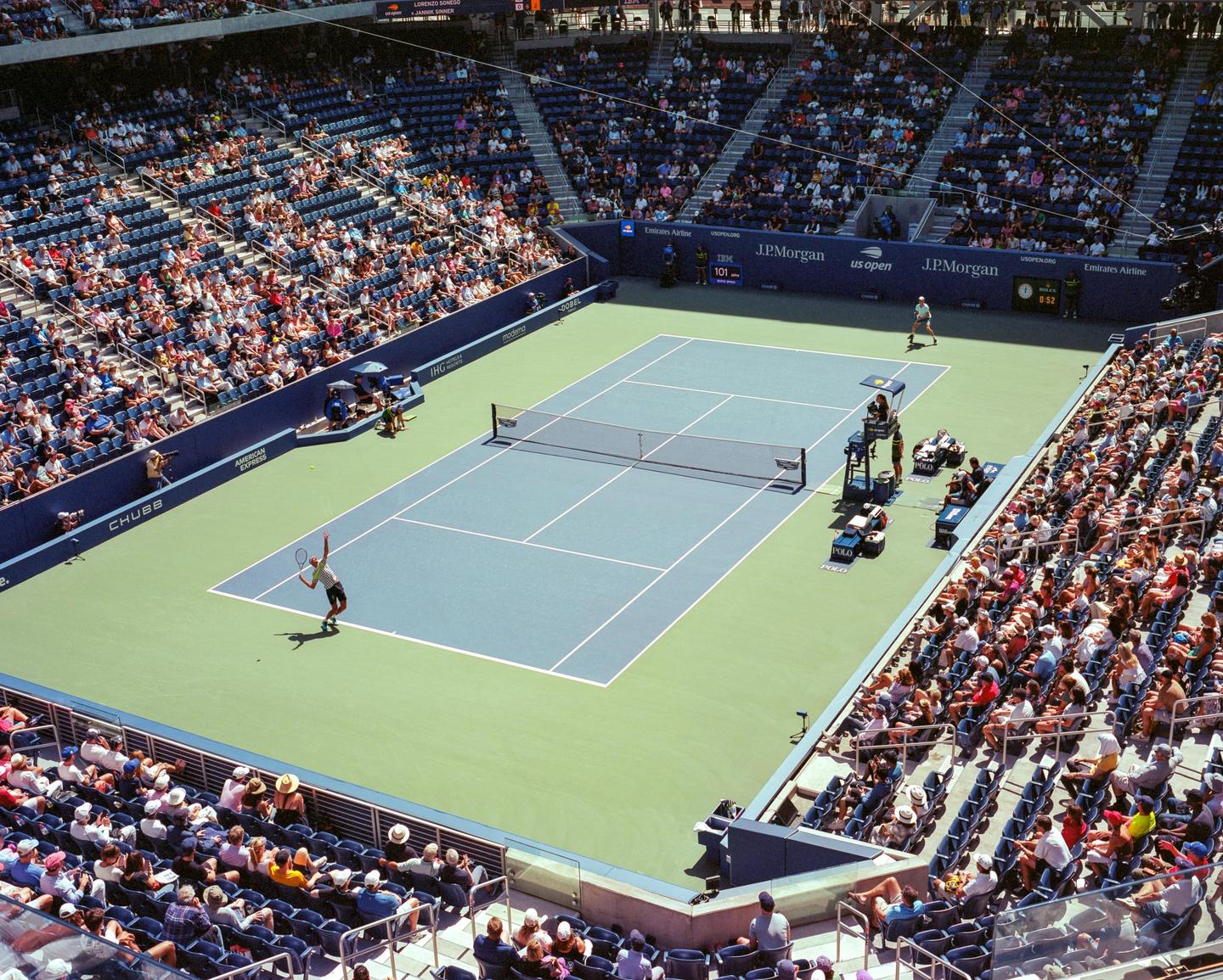 A tennis player serves on a court surrounded by spectators in a large stadium.