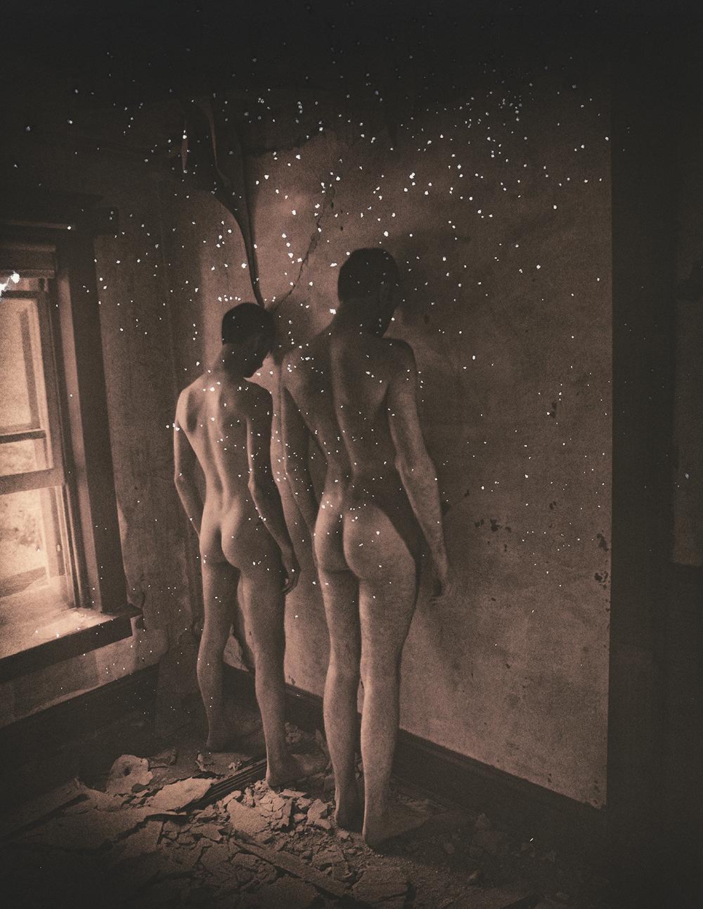Two nude men stand in a derelict room with light particles floating around them.