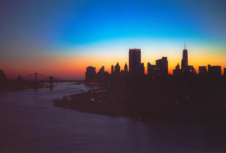 A city skyline and a bridge are silhouetted against a colorful sunset sky over a body of water.