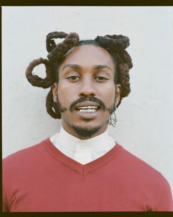 A man with dreadlocks and a beard is wearing a red sweater over a white collared shirt, with a grill on his teeth and an earring.