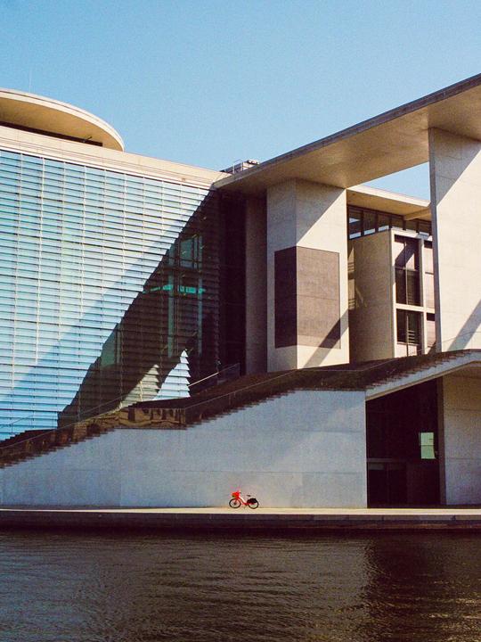 A red bicycle is parked on a concrete walkway next to a body of water, with a modern building and stairs in the background.