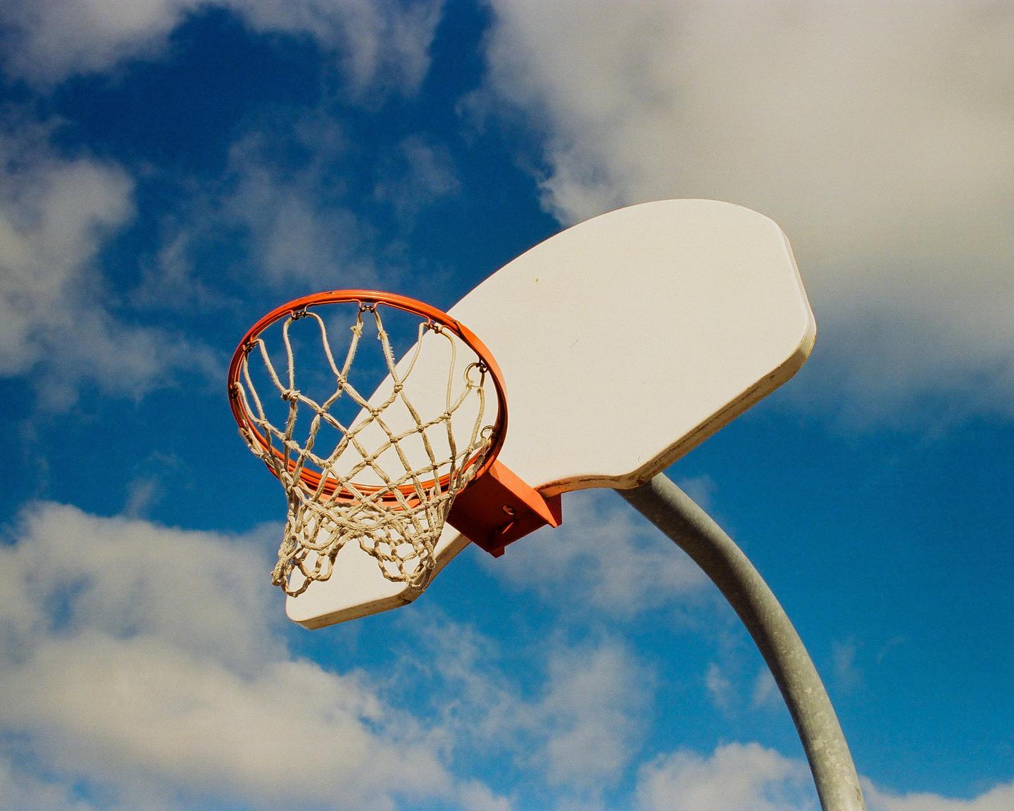 A basketball hoop and backboard are visible against a bright blue sky with scattered clouds.