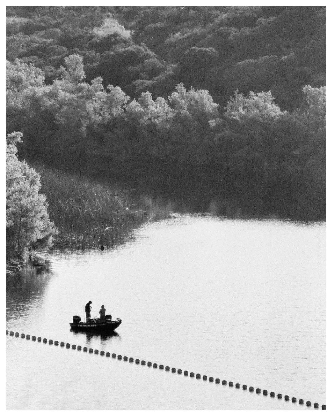 Two people fish from a small boat on a lake with a line of buoys in the foreground and a forested shore in the background.