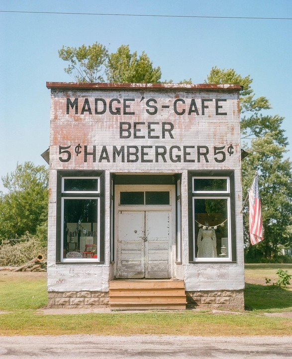 A small, weathered brick building with "MADGE'S-CAFE BEER 5¢ HAMBERGER 5¢" painted on its facade stands beside a dirt road.