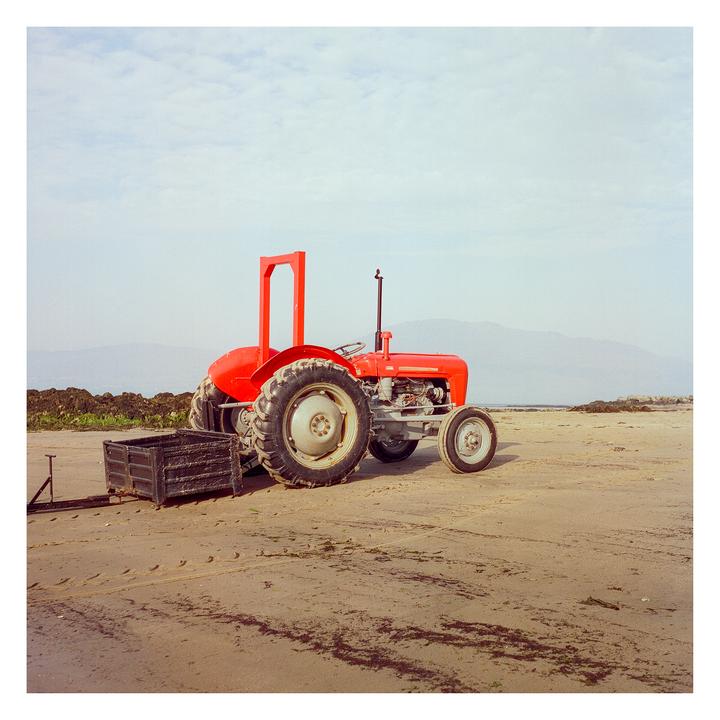 A red tractor is parked on a sandy beach with mountains in the background.