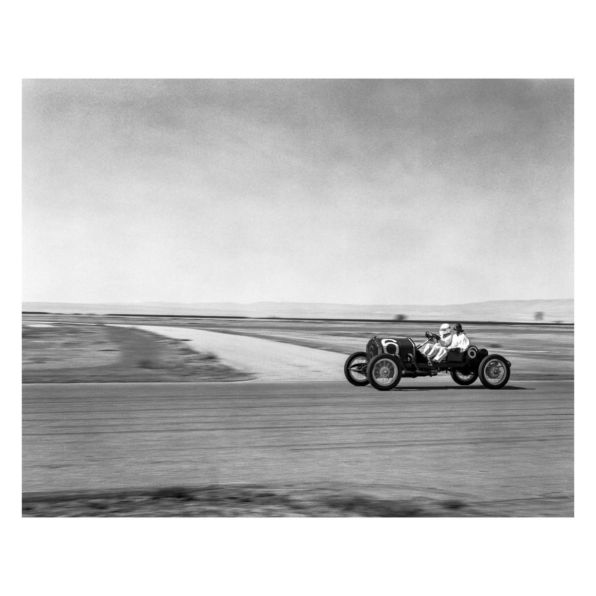 A vintage race car with two men inside speeds along a track under a vast sky.