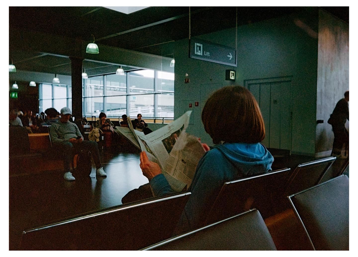 A woman with short brown hair reads a newspaper in a dimly lit waiting area with other people seated around her.