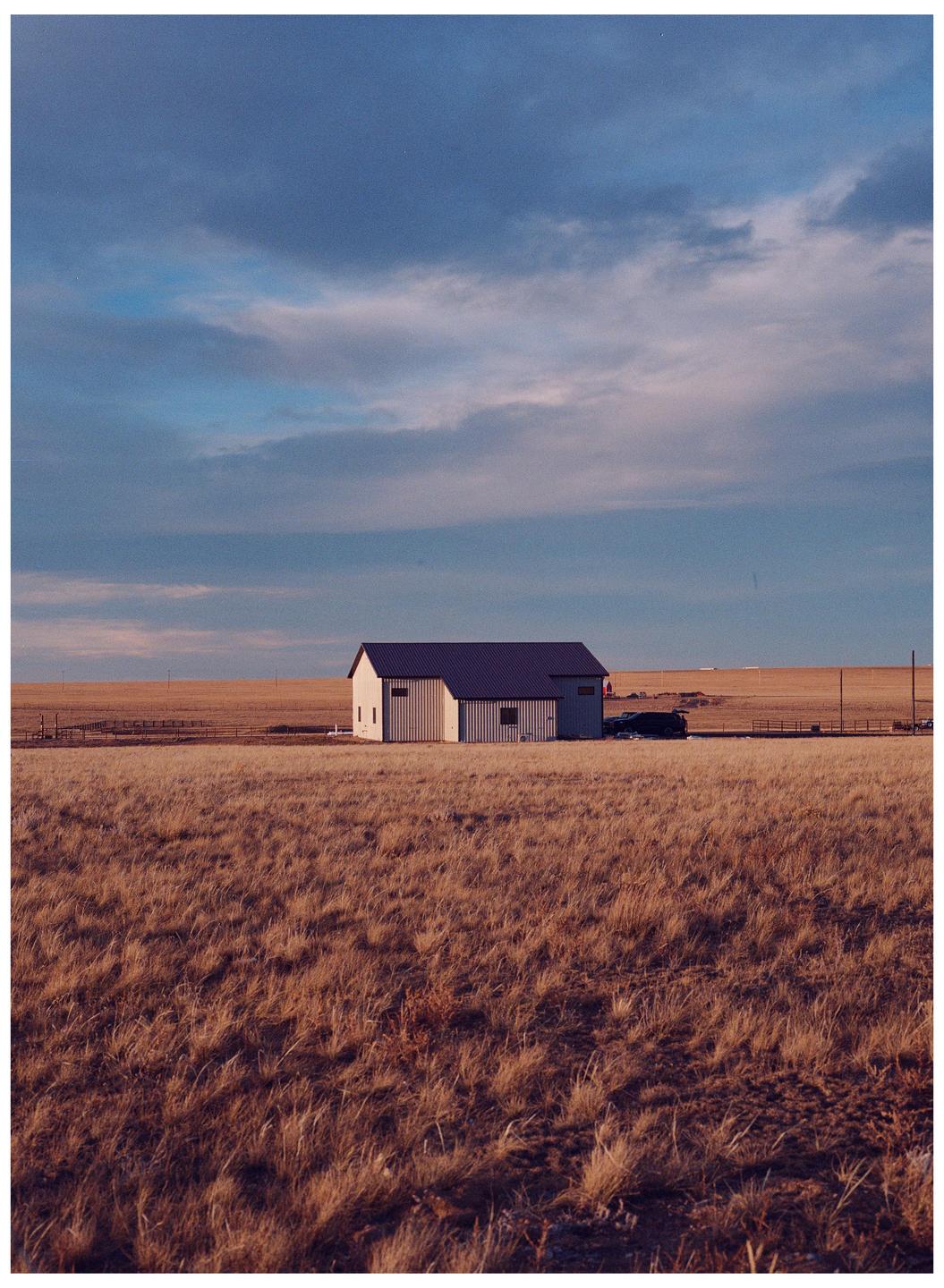 A white barn with a dark roof stands in a field of dry grass under a cloudy sky.