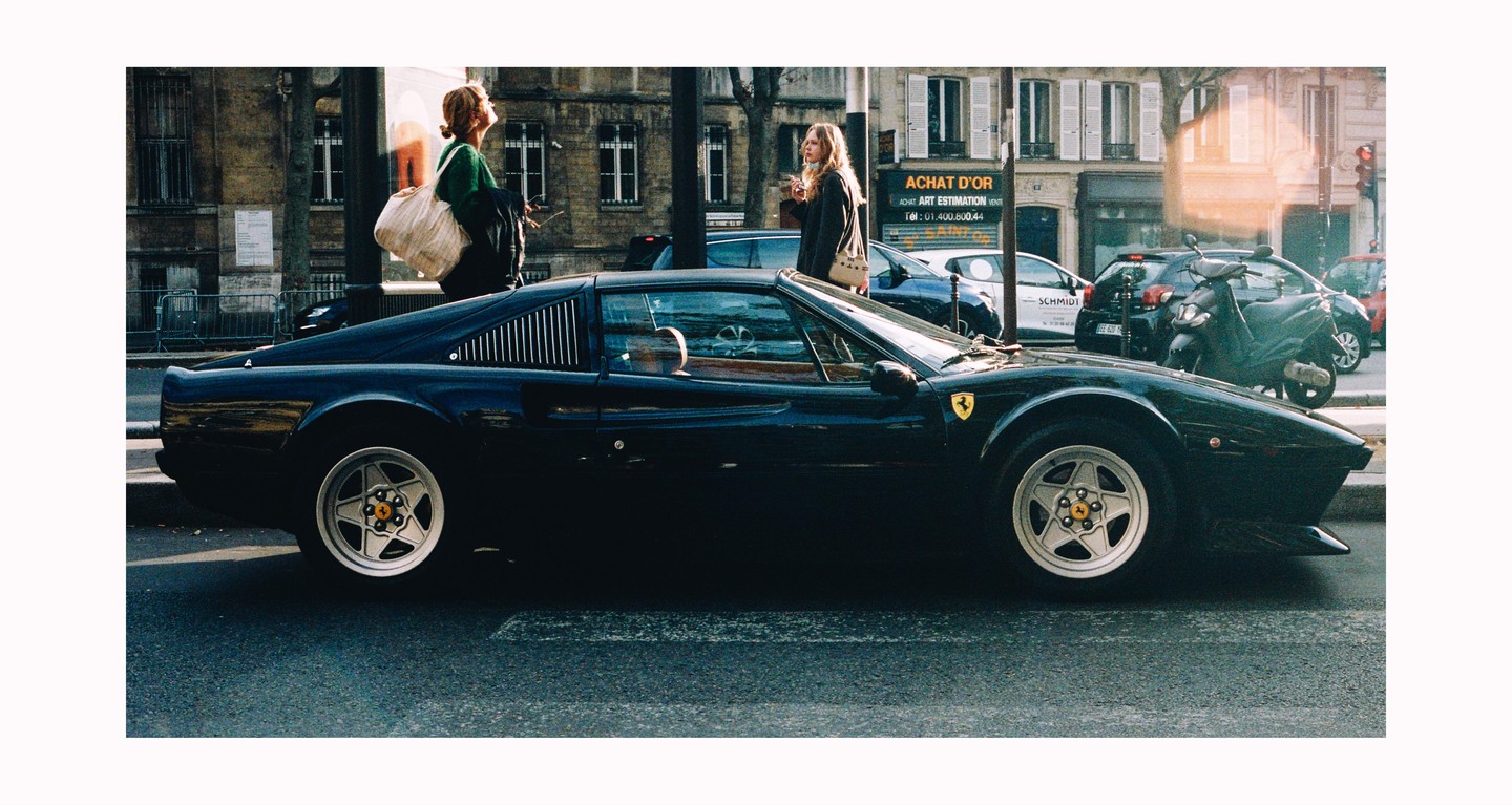 A black sports car is parked on a city street with two women and other vehicles in the background.
