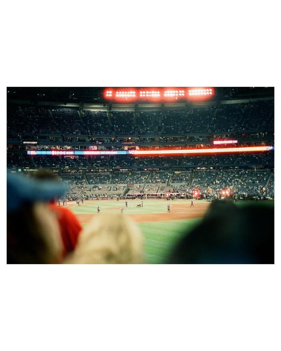 A baseball game is in progress at night, viewed from the stands with blurred heads in the foreground.