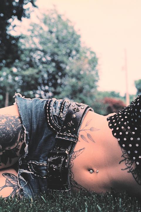 A woman with tattoos and a belly button piercing lies in the grass.