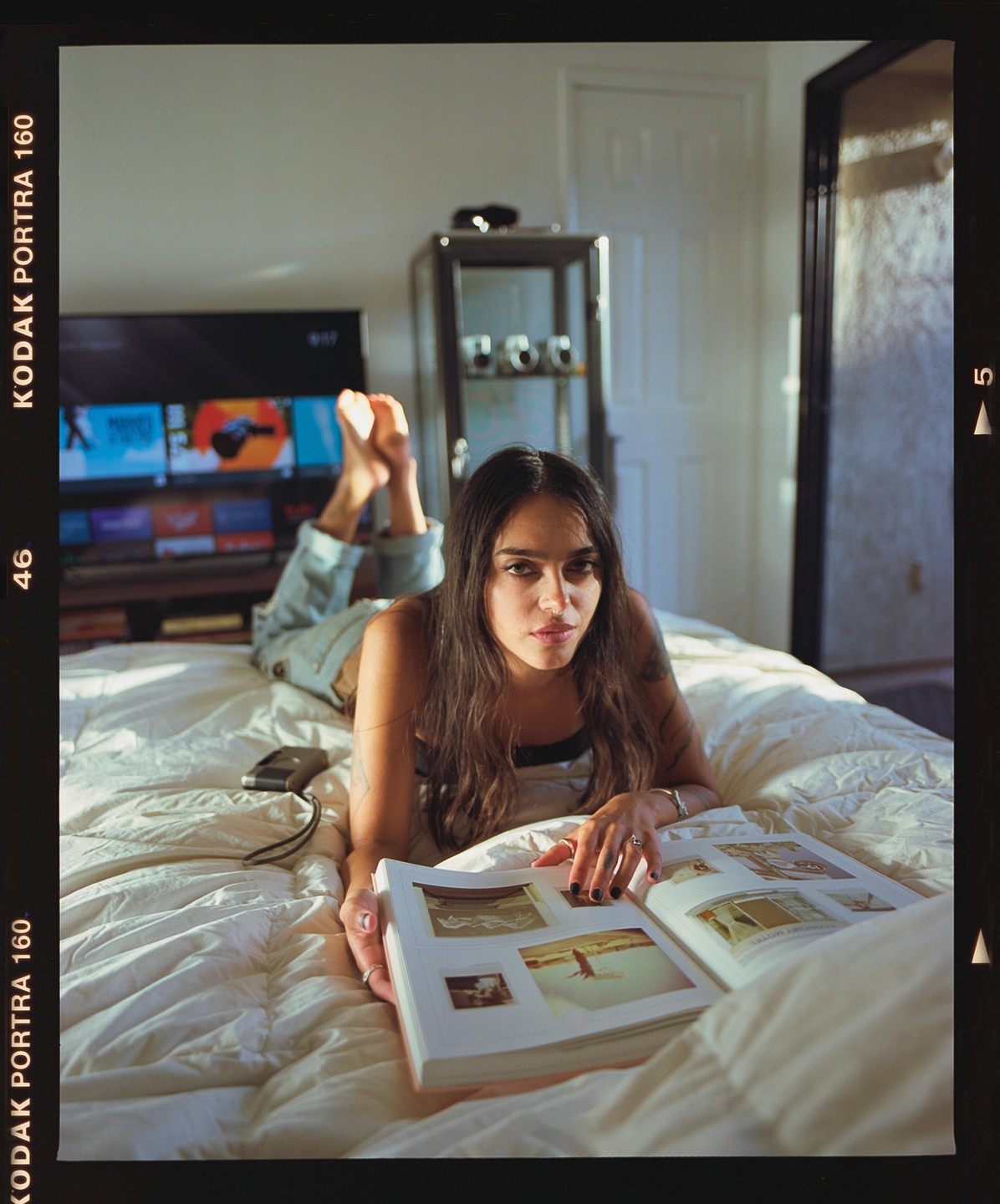 A woman with long hair lies on a bed, looking at a book with images.