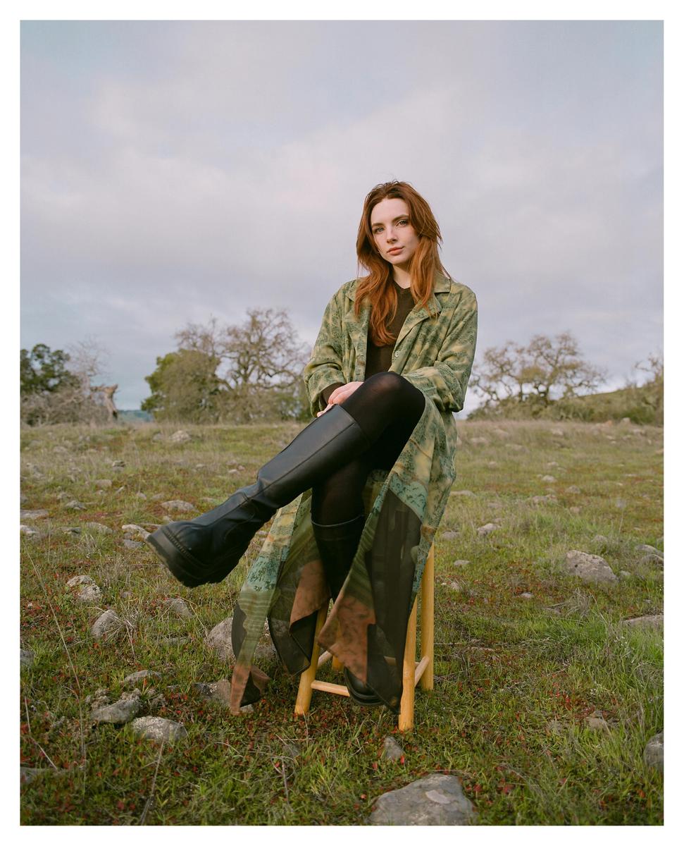 A woman with long red hair sits on a wooden stool in a grassy field.