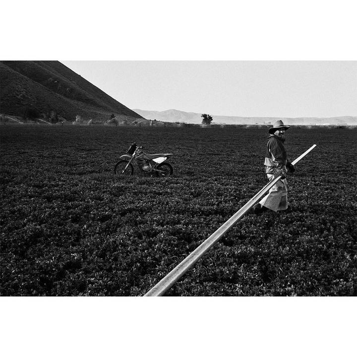 A man in a cowboy hat stands in a field with a motorcycle and a long pipe.