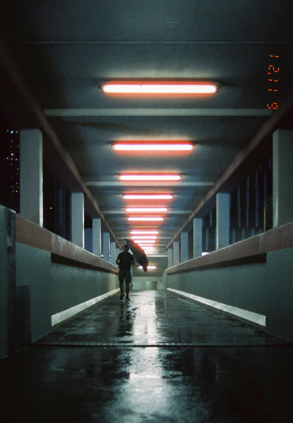 A man walks with an umbrella on a wet walkway illuminated by red fluorescent lights at night.