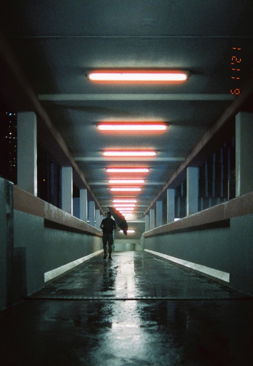A man walks with an umbrella on a wet walkway illuminated by red fluorescent lights at night.